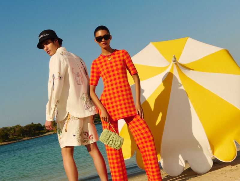 Two models pose on a beach with a yellow-and-white umbrella. One model wears an orange and red checked outfit, the other a patterned shirt and shorts.