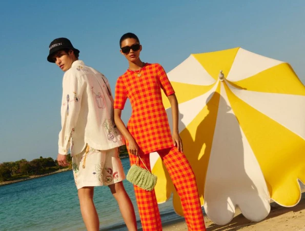 Two models pose on a beach with a yellow-and-white umbrella. One model wears an orange and red checked outfit, the other a patterned shirt and shorts.