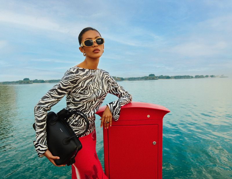 Elegant woman poses outdoors near the water, wearing a zebra-print top, sunglasses, and holding a black handbag next to a red postbox.