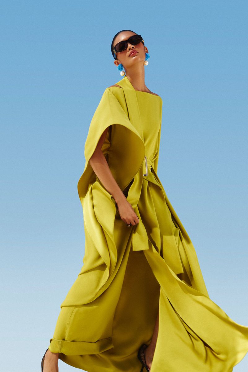 A striking fashion portrait of a woman in a chartreuse gown and sunglasses, standing outdoors against a clear blue sky.