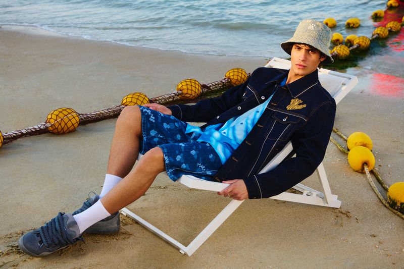 Adult male fashion model poses on beach in bucket hat, denim jacket, and printed shorts, sitting on a white chair near a rope.
