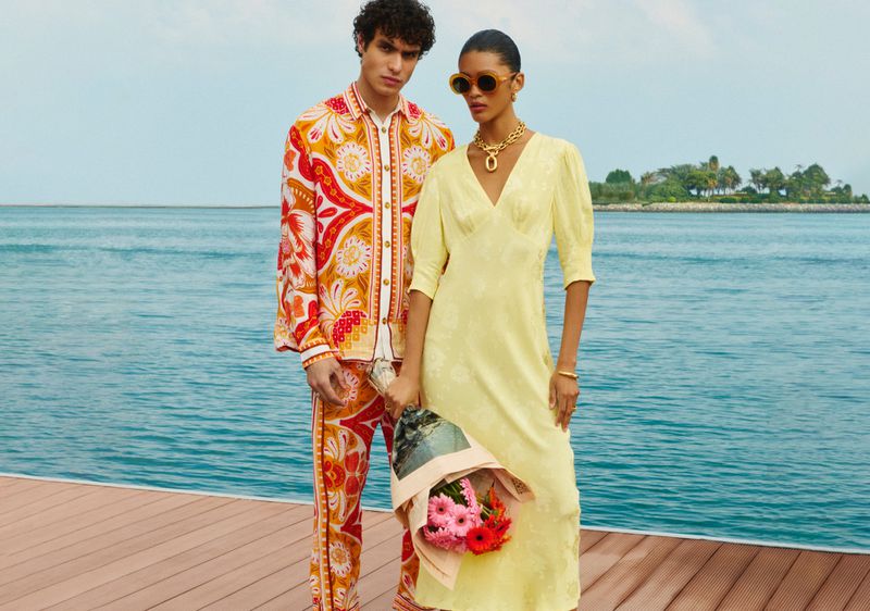 A stylish fashion shot shows a man in a patterned suit and a woman in a yellow dress, standing on a wood dock with flowers and a sea background.
