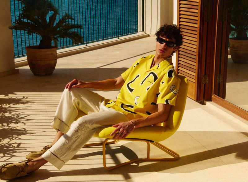Adult male in yellow patterned shirt poses in chair on balcony overlooking ocean, wearing sunglasses, styled cream trousers, and brown shoes.