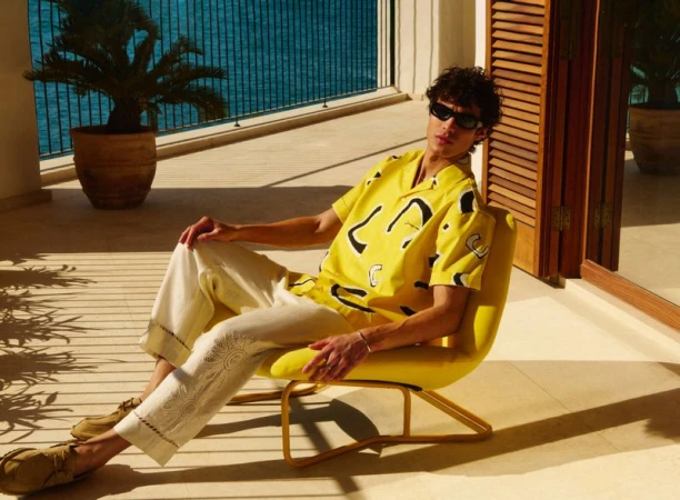 Adult male in yellow patterned shirt poses in chair on balcony overlooking ocean, wearing sunglasses, styled cream trousers, and brown shoes.