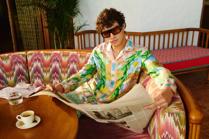 Man in patterned pajamas and sunglasses sits on a patterned sofa outdoors, reading a newspaper, with a coffee cup on a wooden table.