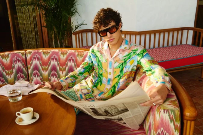 Man in patterned pajamas and sunglasses sits on a patterned sofa outdoors, reading a newspaper, with a coffee cup on a wooden table.