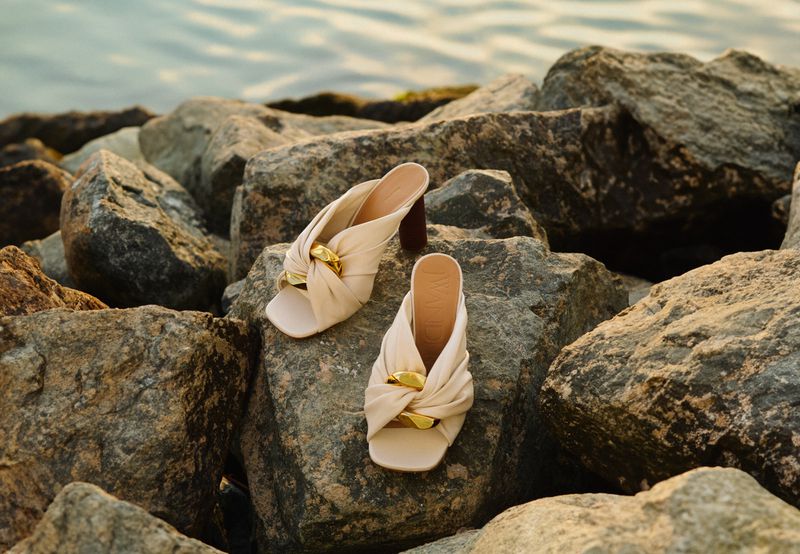 Elegant beige mule shoes with a gold twist detail on rocks near water. Close-up fashion footwear shot, ideal for online commerce.