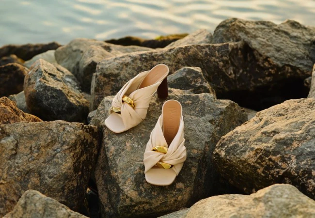 Elegant beige mule shoes with a gold twist detail on rocks near water. Close-up fashion footwear shot, ideal for online commerce.