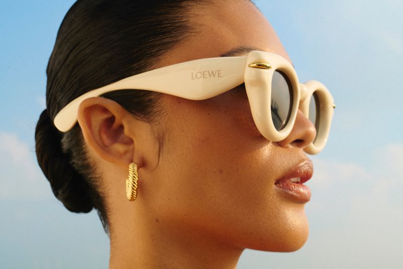 Profile close-up of a woman wearing thick cream sunglasses and a ribbed gold hoop earring against a pale blue sky.