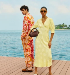 Two models posing on a wooden dock by the water, wearing patterned resort wear and holding a dark handbag.