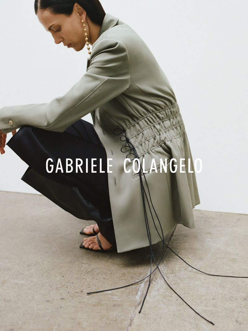 Fashion shot of a woman in a sage blazer and black pants crouching indoors against a light gray backdrop, accessorized with gold earrings.