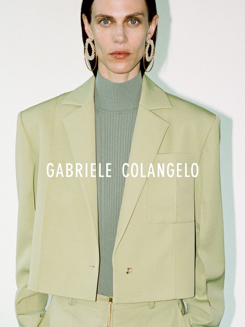 Fashion portrait of a woman with fair skin wearing a sage green blazer, matching pants and earrings, and a light green turtleneck.