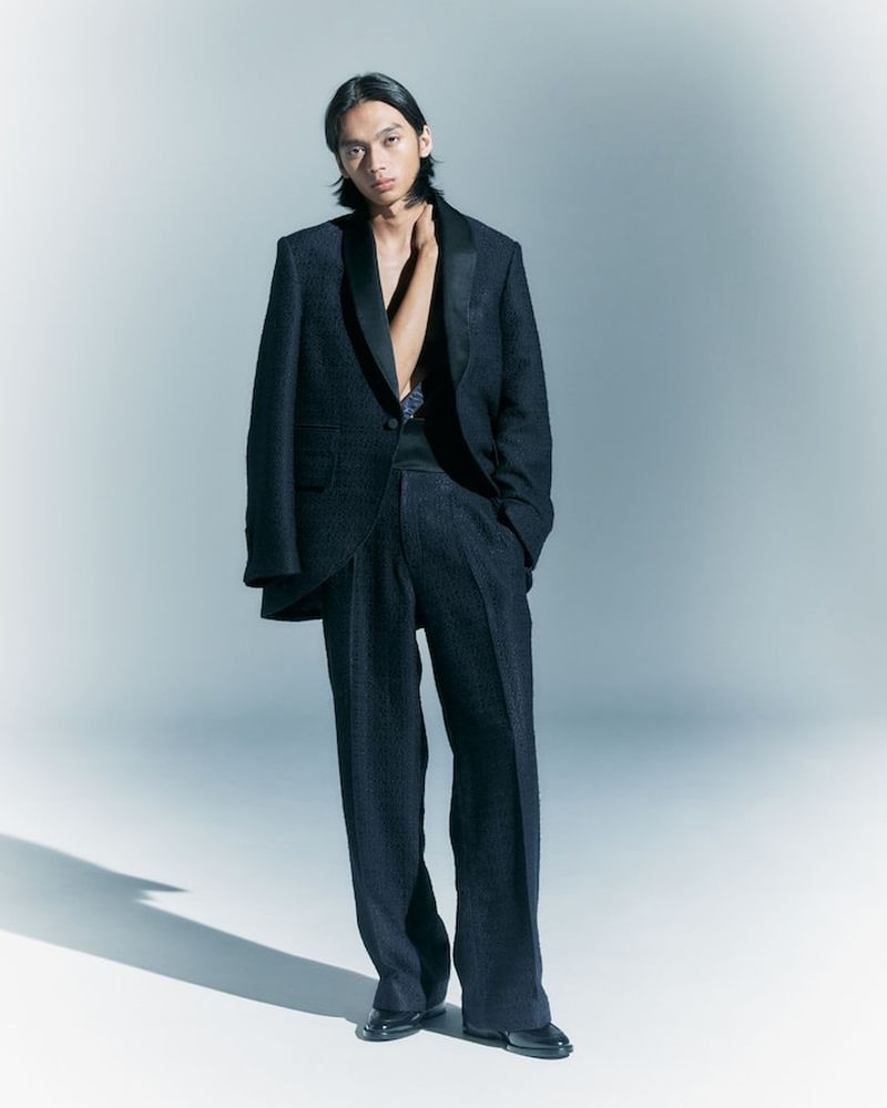 A slender man in a textured dark blue suit poses in a studio with a hand at his neck, looking directly at the camera with a neutral expression.