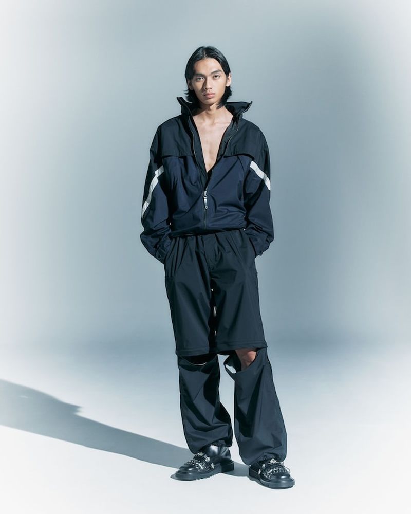 A slender adult male stands in a studio, wearing a navy and white tracksuit jacket with baggy black pants and chunky black shoes.