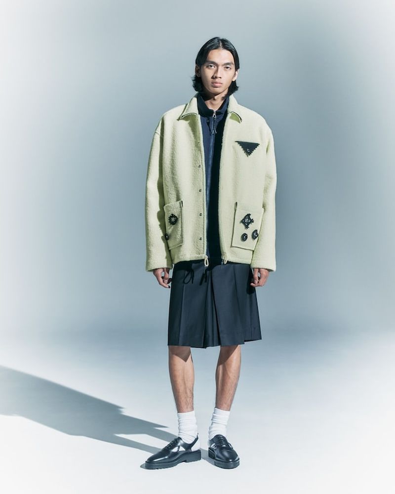 A stylish adult male in a pale yellow jacket and pleated skirt poses in a studio with soft lighting. Fashion editorial style.