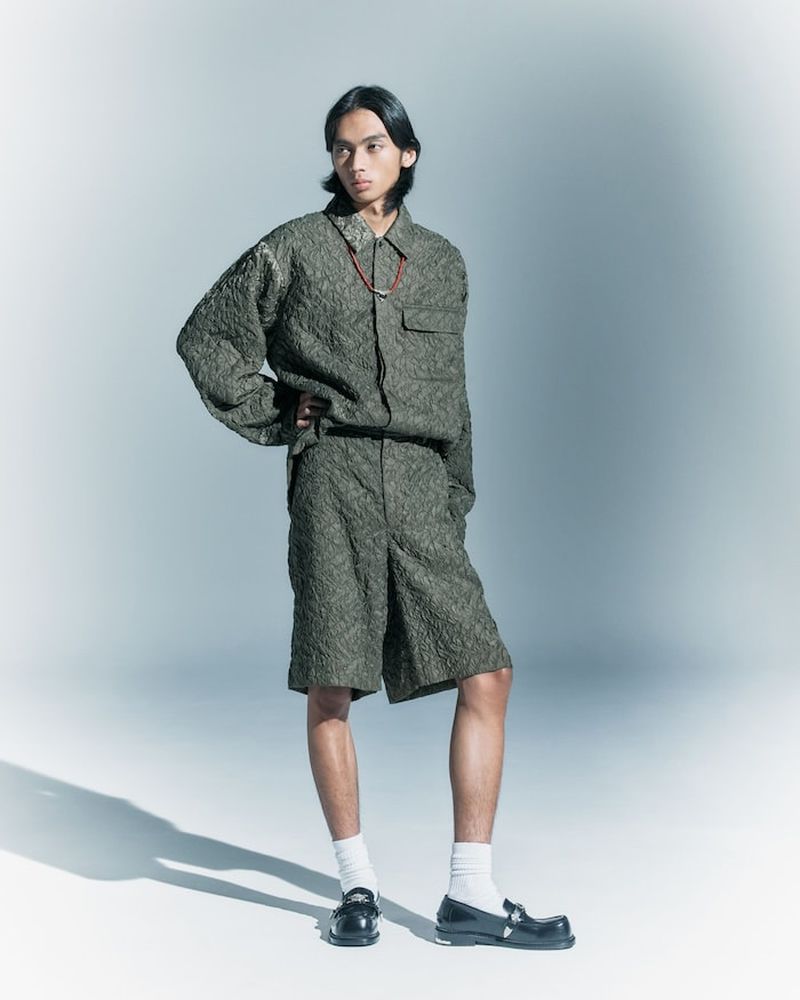 An adult male poses in a photo studio wearing a green textured button-up shirt, shorts, socks, and loafers against a gradient background.