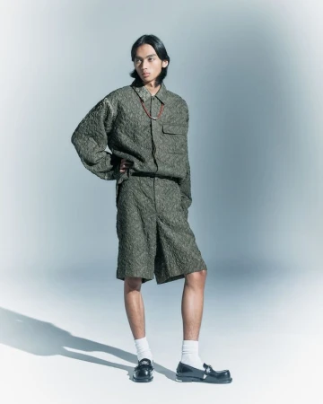 An adult male poses in a photo studio wearing a green textured button-up shirt, shorts, socks, and loafers against a gradient background.