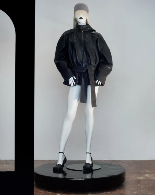 Mannequin in a black leather jacket and grey beanie. The mannequin stands on a glossy black circular stand indoors.