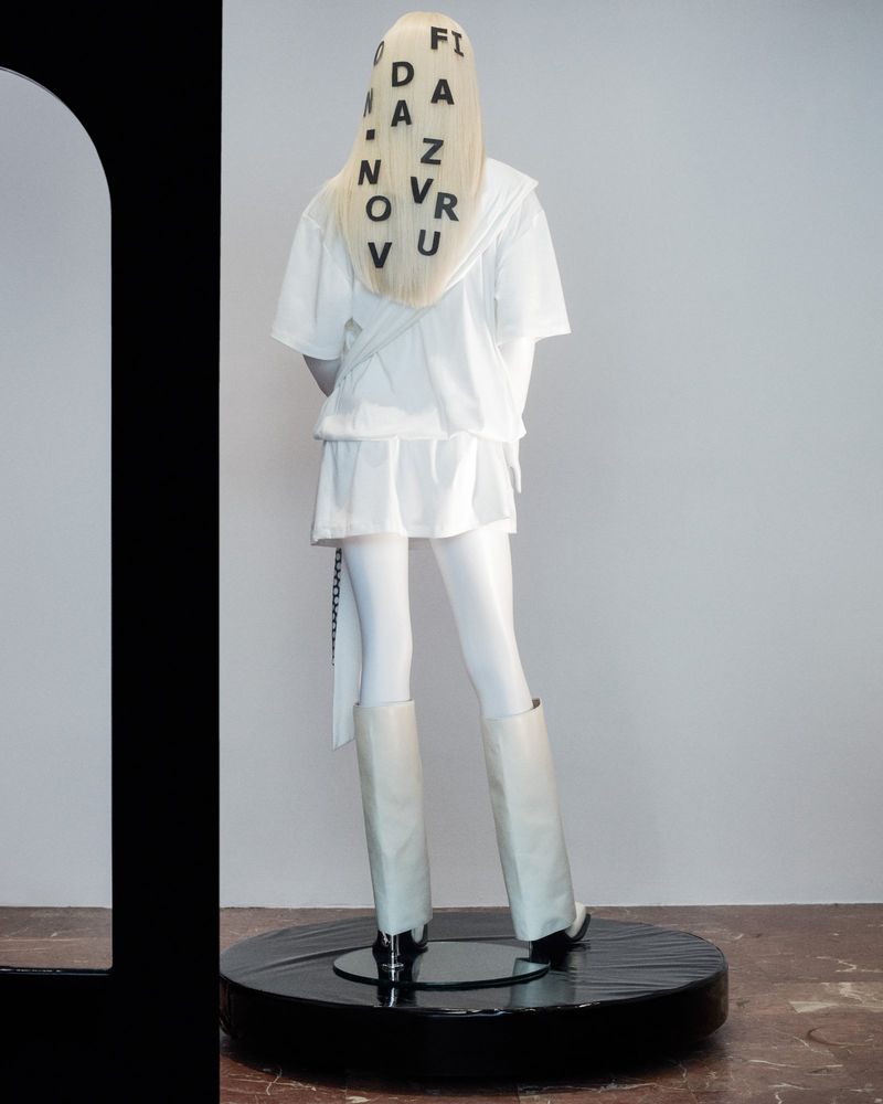 Back view of a fair-skinned woman in a white outfit and white knee-high boots, black letters on her blonde hair, on a black platform.