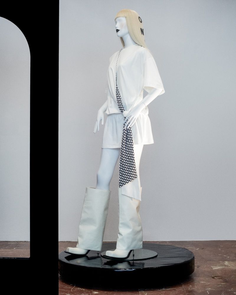 Mannequin in white t-shirt dress and high boots on a black platform. A printed scarf hangs from its neck. Studio setting.