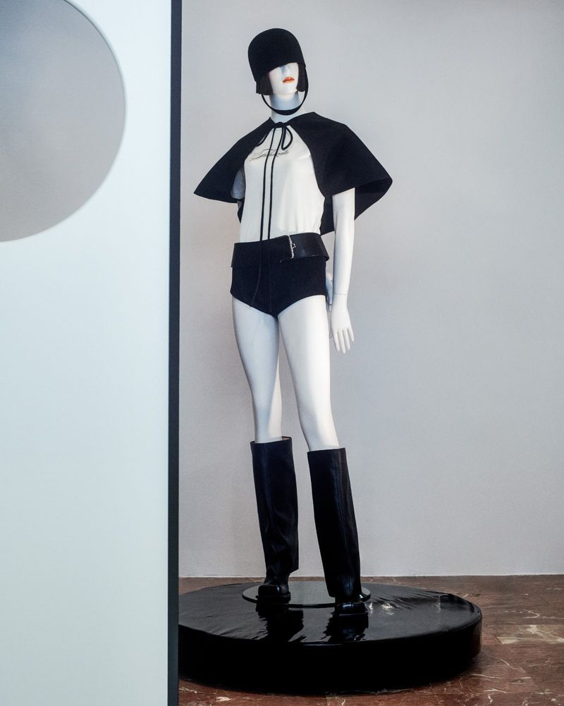 Mannequin in white top, black cape, shorts, hat, and boots stands on a black platform against a white wall in a minimalist display.