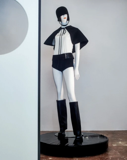 Mannequin in white top, black cape, shorts, hat, and boots stands on a black platform against a white wall in a minimalist display.