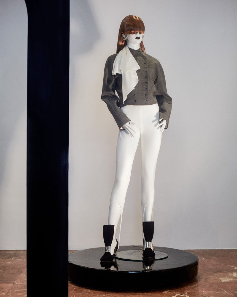 Mannequin stands tall in a gray jacket, white pants, and black boots on a round display platform in a minimalist studio setting.