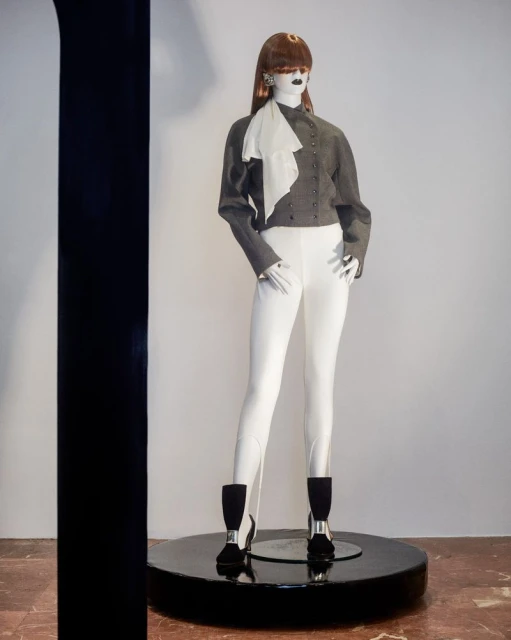 Mannequin stands tall in a gray jacket, white pants, and black boots on a round display platform in a minimalist studio setting.