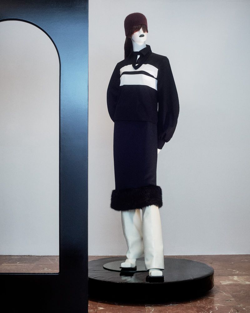 Fashion mannequin wearing a black and white striped shirt with black skirt and white boots, posing near a modern black arch.