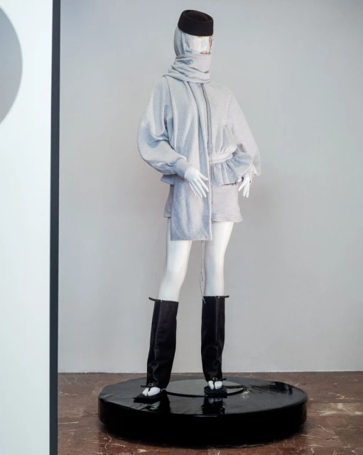 Mannequin wearing a light grey hooded outfit with a matching scarf, black boots, and a black beanie on a black pedestal.