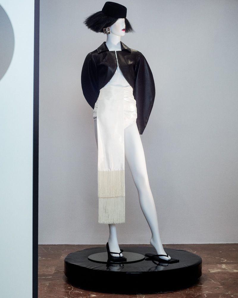 Fashion mannequin in white and black ensemble, featuring a short black jacket, dress with fringes, wide brim hat, and black heeled sandals.