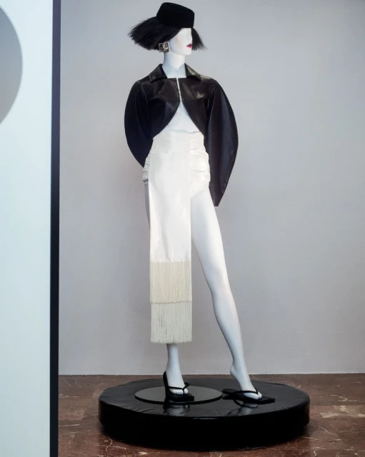 Fashion mannequin in white and black ensemble, featuring a short black jacket, dress with fringes, wide brim hat, and black heeled sandals.