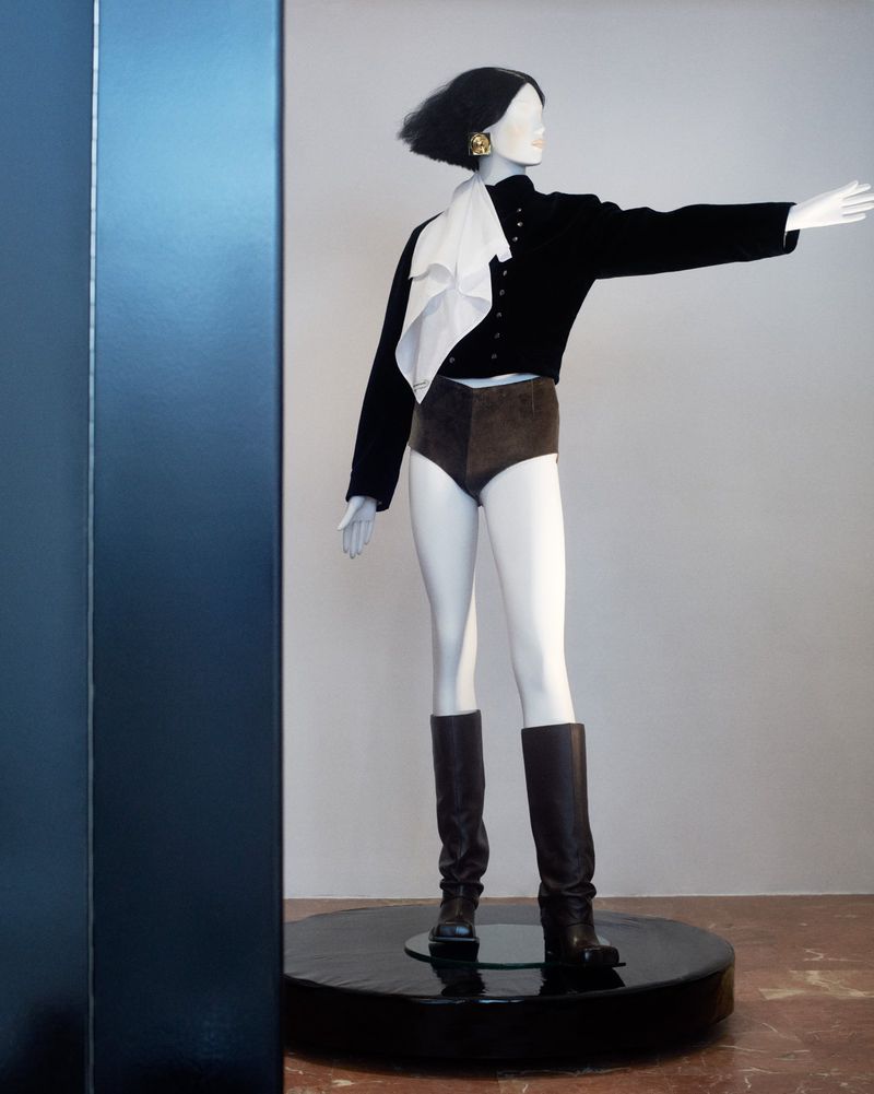 Mannequin displays black cropped jacket, brown high-waisted shorts, and dark boots. White scarf and gold earrings add accent in studio.