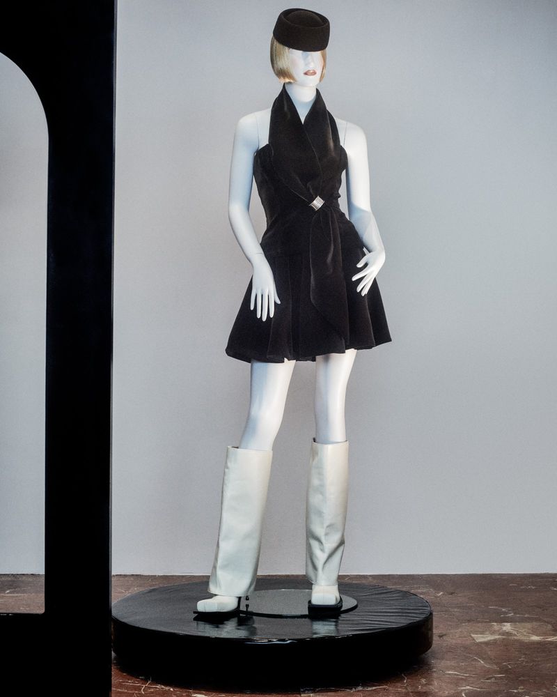Mannequin wearing a dark halter dress with a silver buckle and white boots, topped with a black hat against a white background.