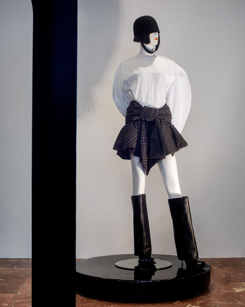 Mannequin displays a fashion ensemble featuring a white blouse, plaid skirt-wrap, helmet, and black boots, set against a simple background.
