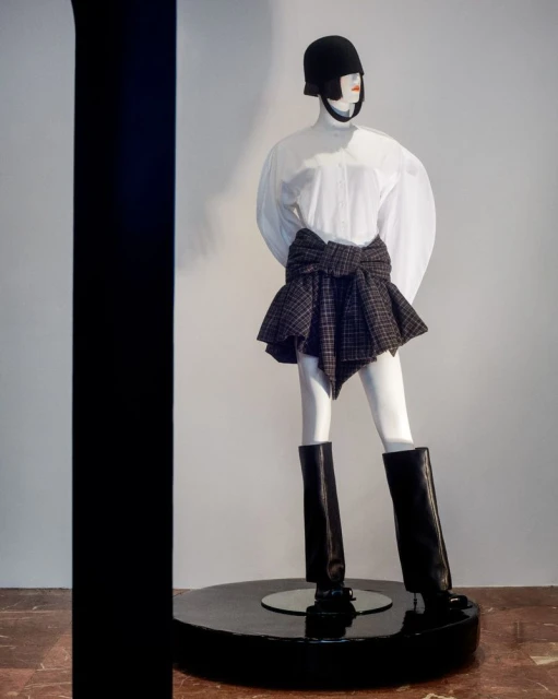 Mannequin displays a fashion ensemble featuring a white blouse, plaid skirt-wrap, helmet, and black boots, set against a simple background.
