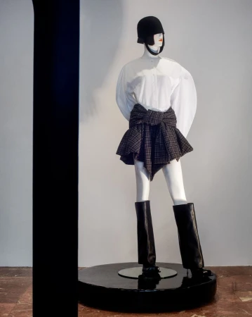 Mannequin displays a fashion ensemble featuring a white blouse, plaid skirt-wrap, helmet, and black boots, set against a simple background.