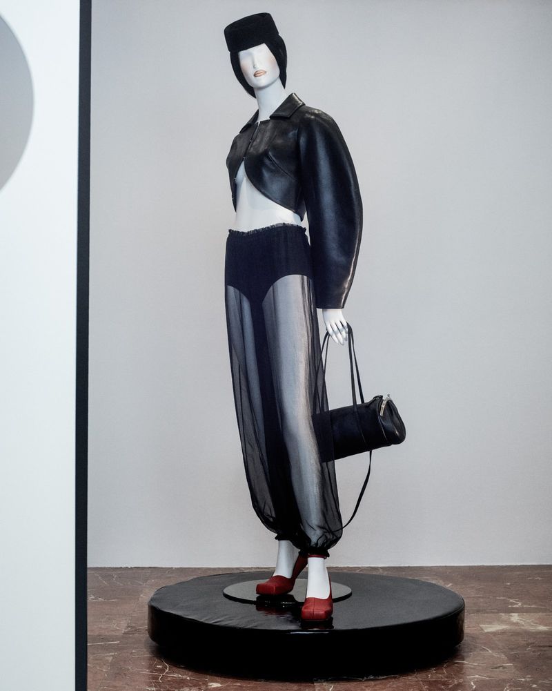 Mannequin displaying a black faux leather jacket, sheer pants, and red heels on a black platform against a neutral background.