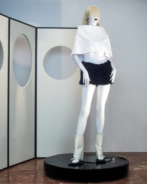 Mannequin displays a fashion ensemble including a white top, fur shorts, and white boots on a black platform, set against a geometric backdrop.