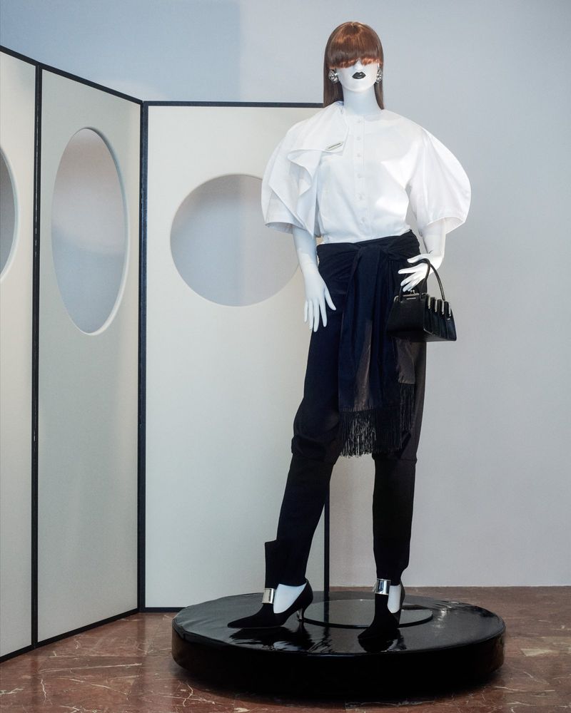 Mannequin displays a white blouse with oversized sleeves, black pants and pumps, and a small black handbag in a minimalist setting.