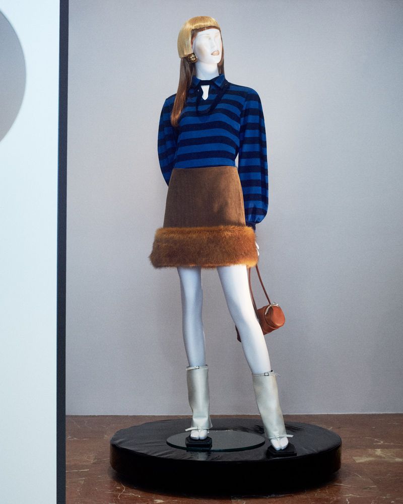 Mannequin displays blue striped long-sleeved shirt with brown skirt, furry trim, white boots, and matching purse on a black display.
