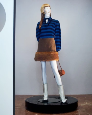 Mannequin displays blue striped long-sleeved shirt with brown skirt, furry trim, white boots, and matching purse on a black display.