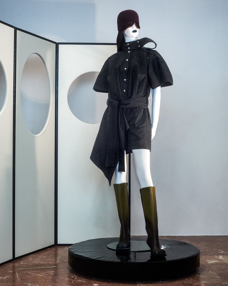 Mannequin displays a modern short black dress with a matching belt, knee-high dark green boots, and a dark red beanie against a white background.