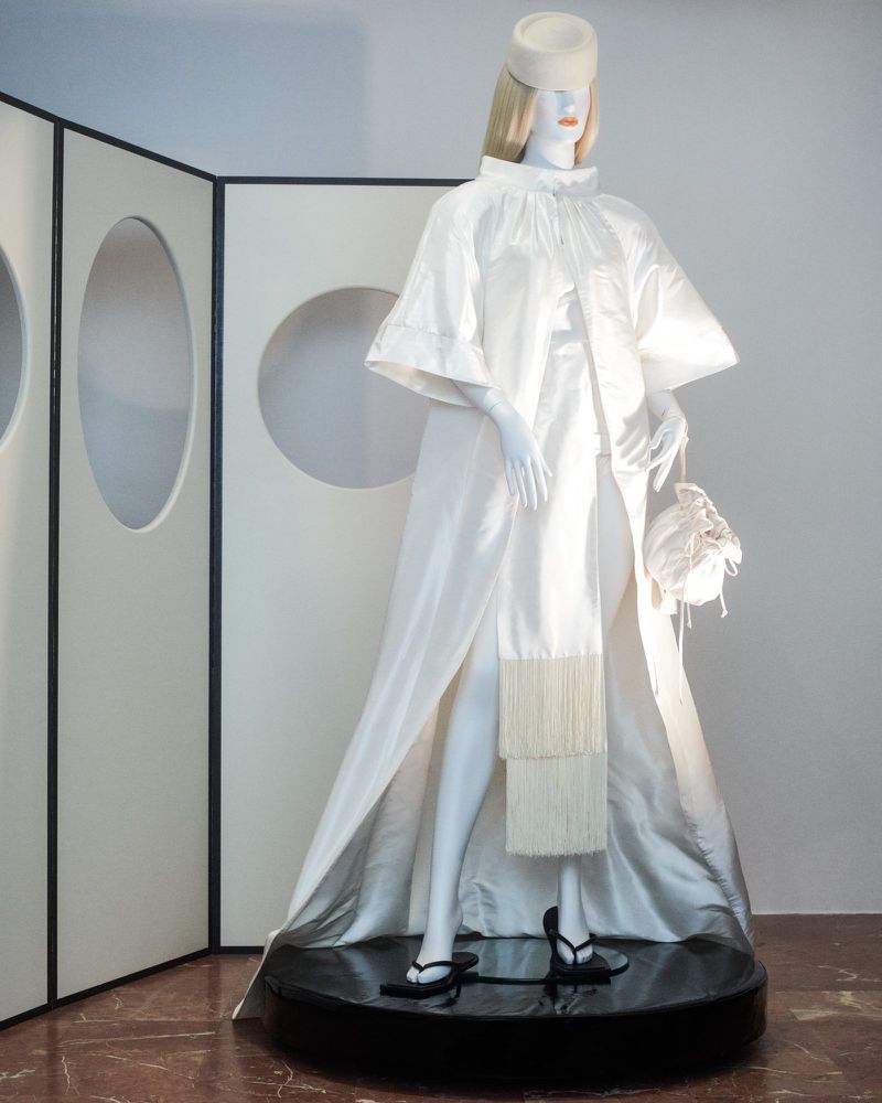 Mannequin displays white couture dress with hat and handbag, standing on a glossy black platform in front of a modern display screen.