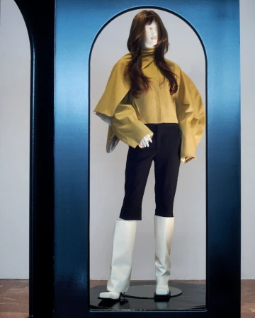 Mannequin displays olive-green top, black trousers, and white boots in a dark display case against a white background.