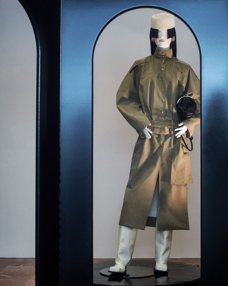 Mannequin displays an olive trench coat, white hat, and handbag within a sleek, modern black display case in an indoor setting.