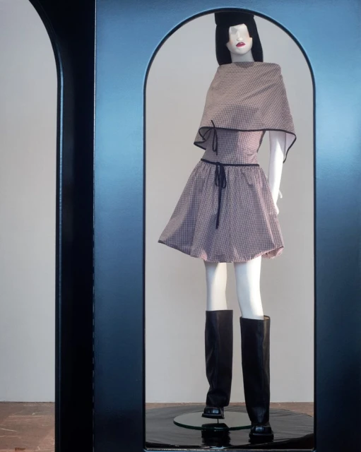 Mannequin displays a retro-style pink gingham dress and cape, complemented by black knee-high boots, framed by a black archway.