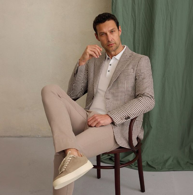 Adult male sitting on a chair wearing a blazer, shirt, and trousers. Indoor shot with green and beige background.