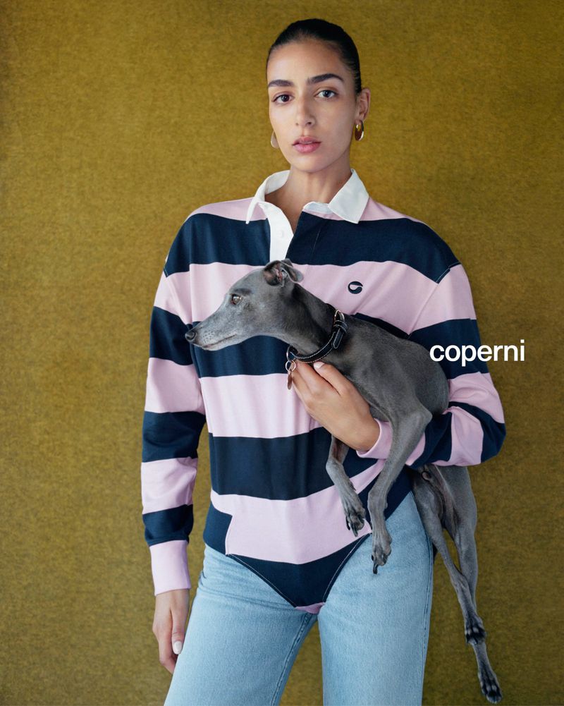 Adult female poses indoors with a grey Italian Greyhound dog, wearing a pink and navy striped shirt and light blue jeans against an olive backdrop.