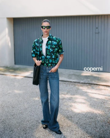 Woman in floral shirt, jeans, and sunglasses stands on a paved road in daylight with a tote bag and building in the background.